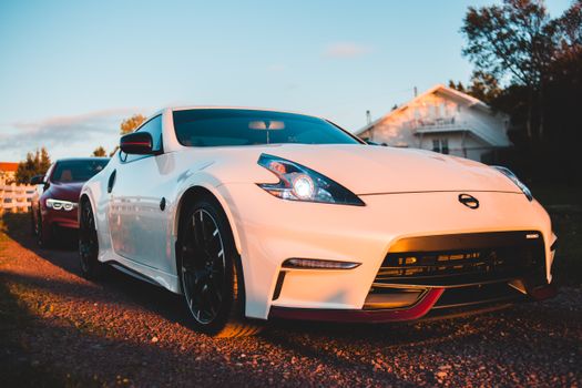 Luxury white sports car with glowing headlights parked on rough road near small houses against blue sky in countryside