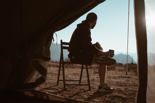一名男子在帐篷外喝着咖啡，俯瞰日落时分的群山，享受宁静的时刻