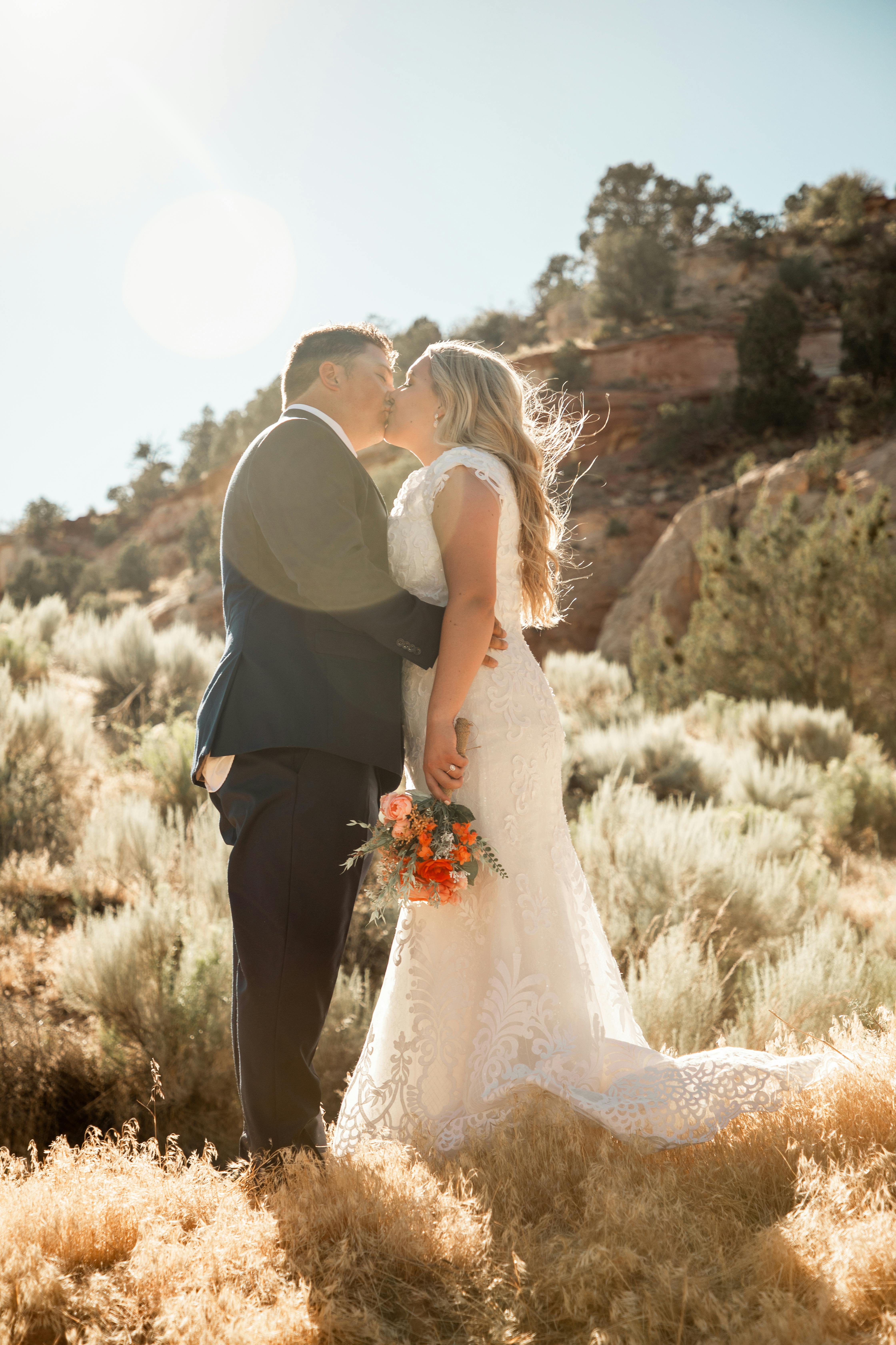 A couple shares a romantic kiss in a sunlit outdoor wedding setting.