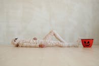 一个穿着木乃伊服装的孩子躺在室内，手里拿着万圣节水桶，非常适合季节性主题