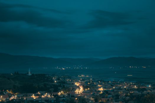 Illuminated village under a moody night sky in Romania, showcasing a serene blend of architecture and nature.