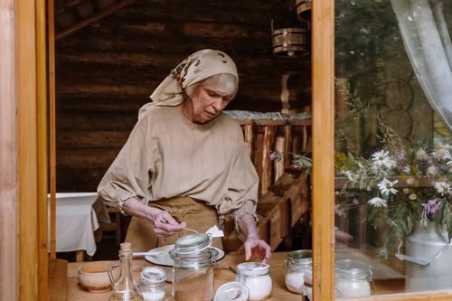 Free Elderly woman preparing food in a rustic kitchen setting, mixing ingredients with jars surrounding her. Stock Photo