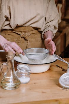 A woman sifts flour in a rustic kitchen, emphasizing homemade cooking and preparation.