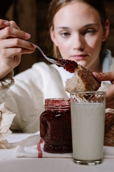 Close-up of a woman spreading jam on bread with a glass of milk on a breakfast table.