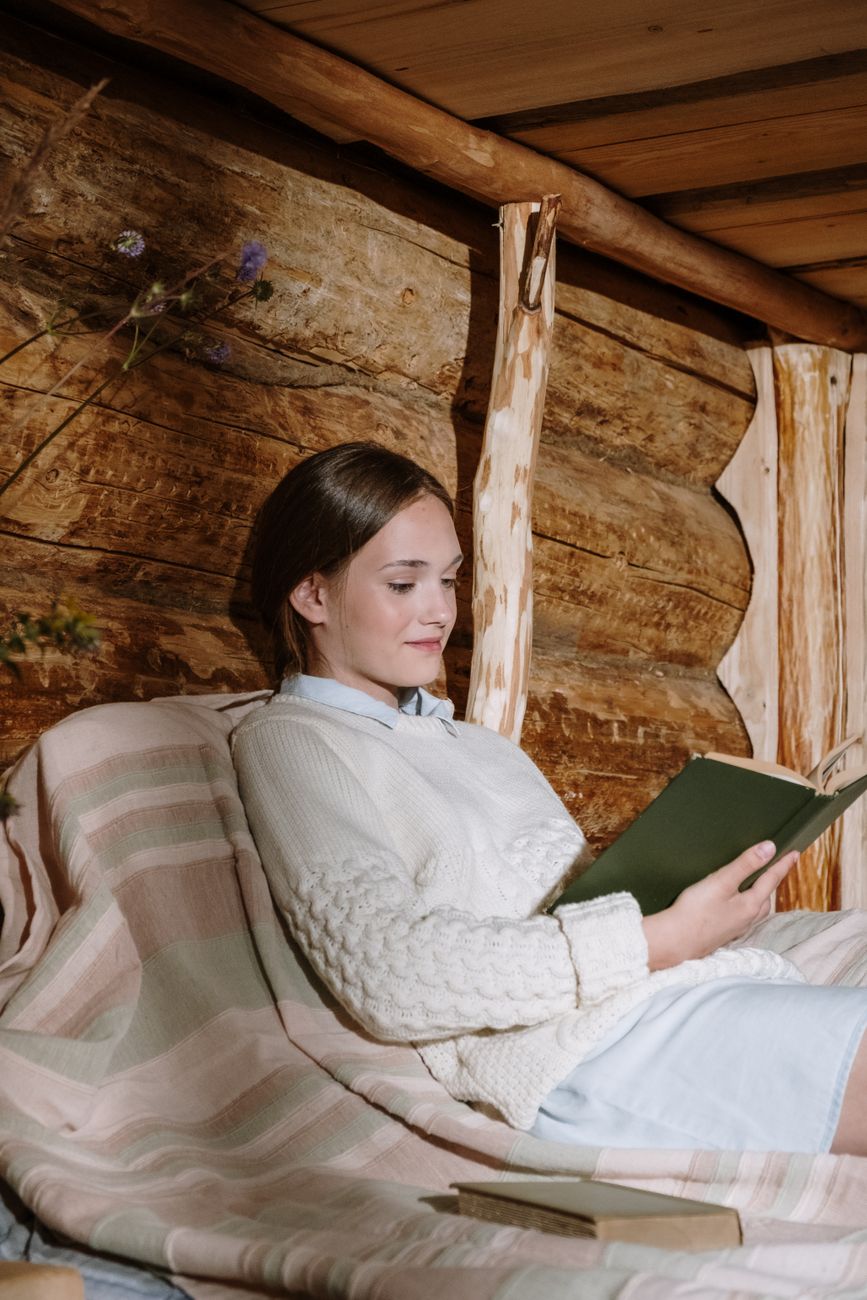 Young woman reading a book in a cozy, wooden cabin setting, wrapped in a knitted sweater.