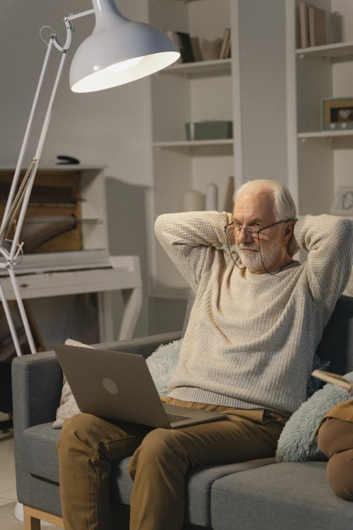 A Man in Beige Knitted Sweater Sitting on the Couch while Using Laptop