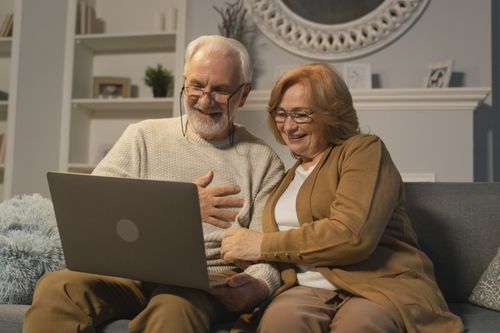 Free Happy senior couple sitting on a sofa having a joyful video call on a laptop in a cozy living room. Stock Photo