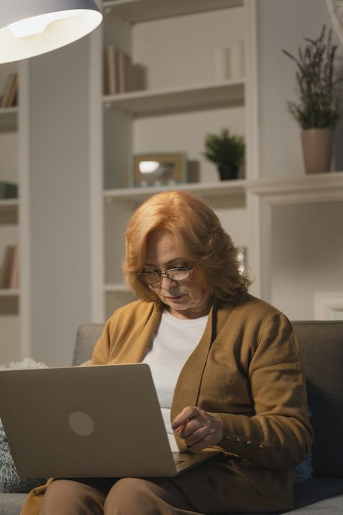 Free A senior woman with glasses sits on a sofa using a laptop in a cozy living room. Stock Photo