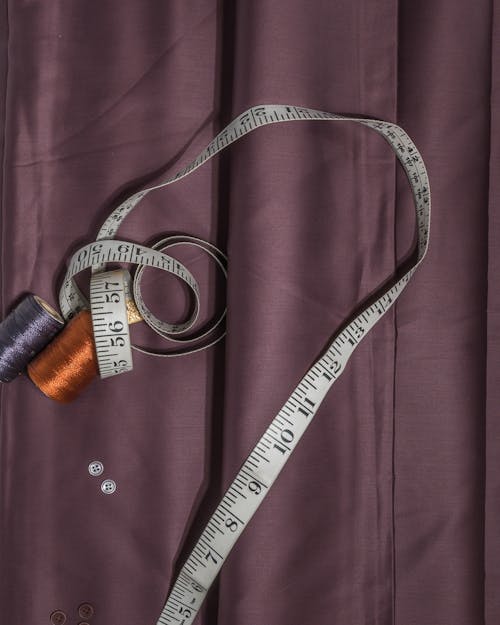 High angle composition of colorful spools of thread located on unwound measuring tape and near small buttons on violet textile