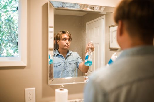 Man cleaning bathroom mirror with blue spray bottle reflection in the frame.