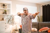 A man in a checkered shirt and cap entertains with a mop in a cozy living room.