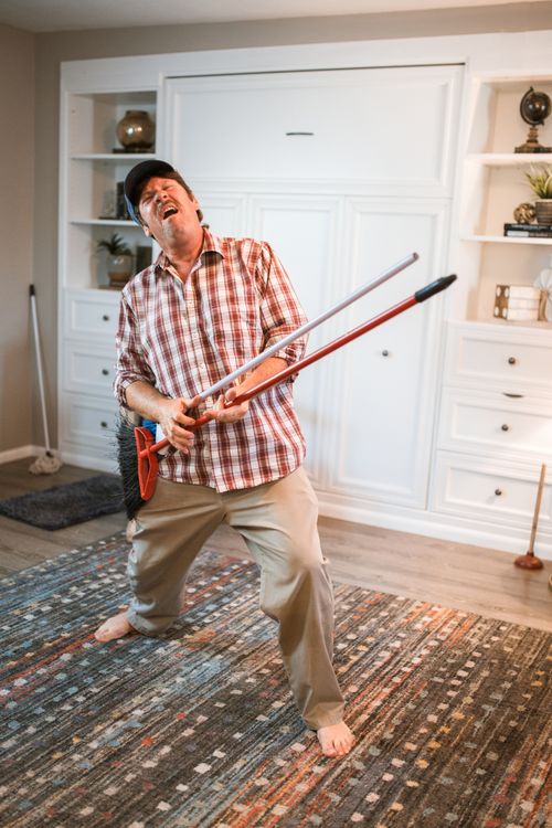 Free A man in a checkered shirt rocking out with a broom as a guitar in a cozy indoor setting. Stock Photo