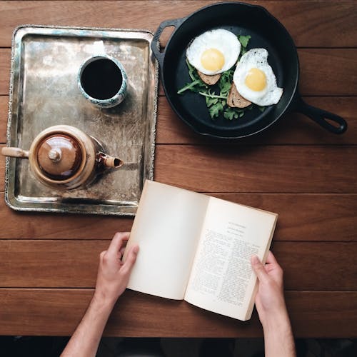 Free Crop person at table with book and breakfast Stock Photo