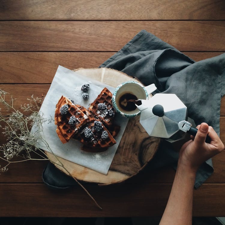 Free From above of crop anonymous person holding pot and pouring coffee in mug near fresh waffles with berries Stock Photo