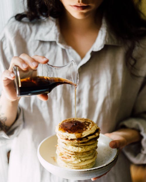 Free Crop faceless female in white shirt holding plate with delicious homemade pancakes and pouring syrup Stock Photo