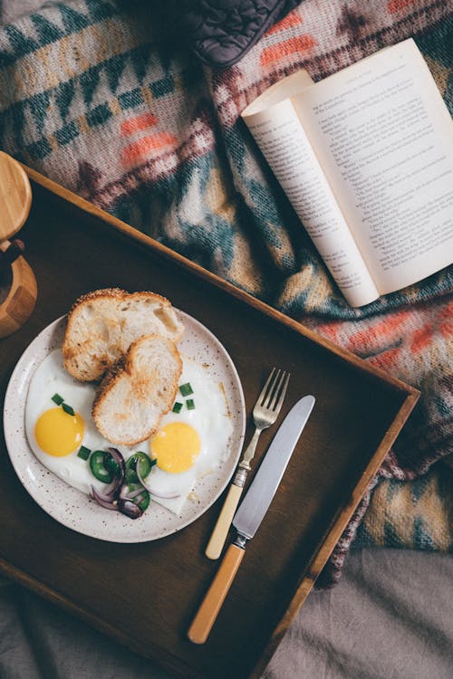 Free From above of opened book on bed near wooden tray with delicious breakfast from fried eggs and onion Stock Photo