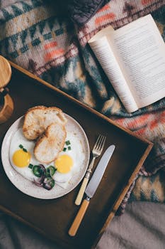 From above of opened book on bed near wooden tray with delicious breakfast from fried eggs and onion