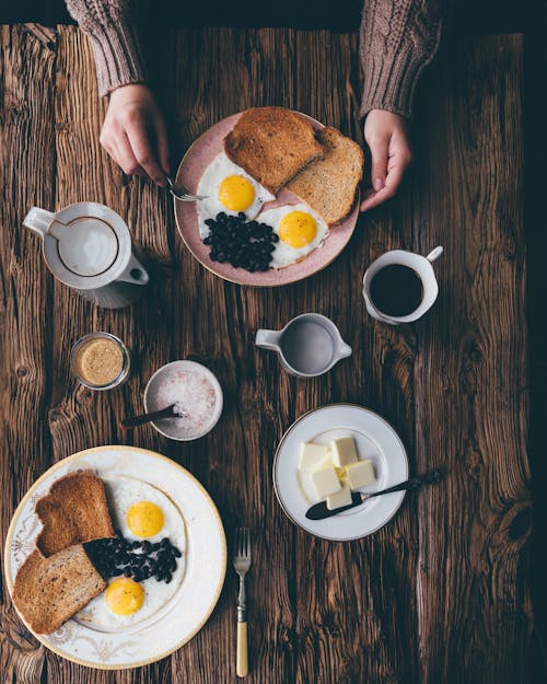Free From above of crop anonymous person in sweater holding fork and plate with delicious breakfast on table with cup of coffee and butter Stock Photo