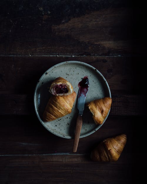 From above of fresh baked croissants with jam in ceramic plate near knife on wooden table