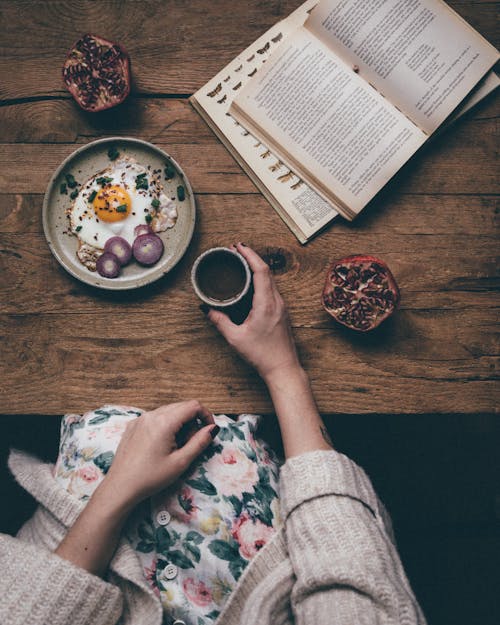 Free From above of crop unrecognizable female holding cup of hot coffee at table with fresh breakfast and pomegranate while reading Stock Photo