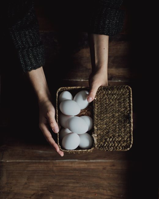 From above of crop unrecognizable person in sweater holding straw basket full of chicken eggs on wooden table