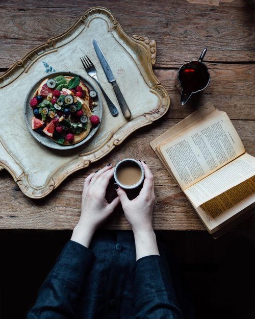 Free Crop faceless female holding cup of hot aromatic coffee on table near meal while reading book Stock Photo