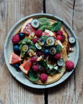 Delicious pancakes topped with fresh berries, kiwi, and mint on a rustic wooden table.