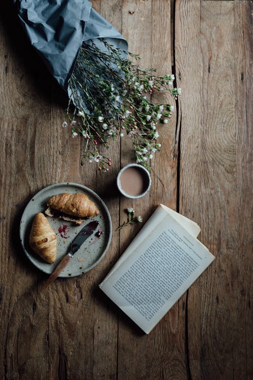 Free Top view flat lay of cup of coffee with croissant served on plate place on wooden table near opened book and wildflowers Stock Photo