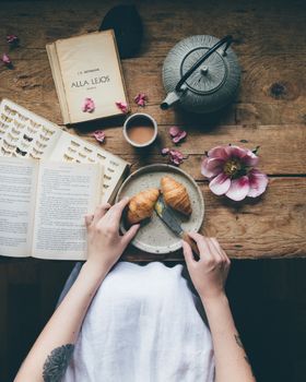Top view of anonymous female standing near wooden table with coffee and opened books near kettle while eating croissants served on plate on breakfast