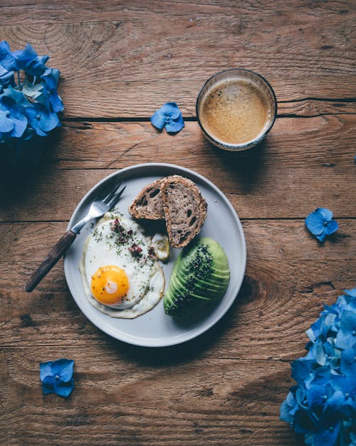 Top view of fried egg with slices of avocado and bred served on plate on wooden table near hot coffee and blue flowers