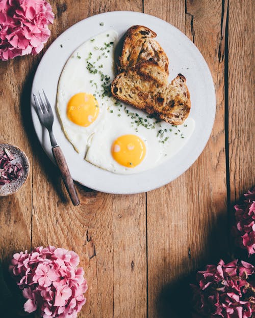 From above of appetizing fried eggs served on white plate with bread placed on wooden table with pink flowers in morning