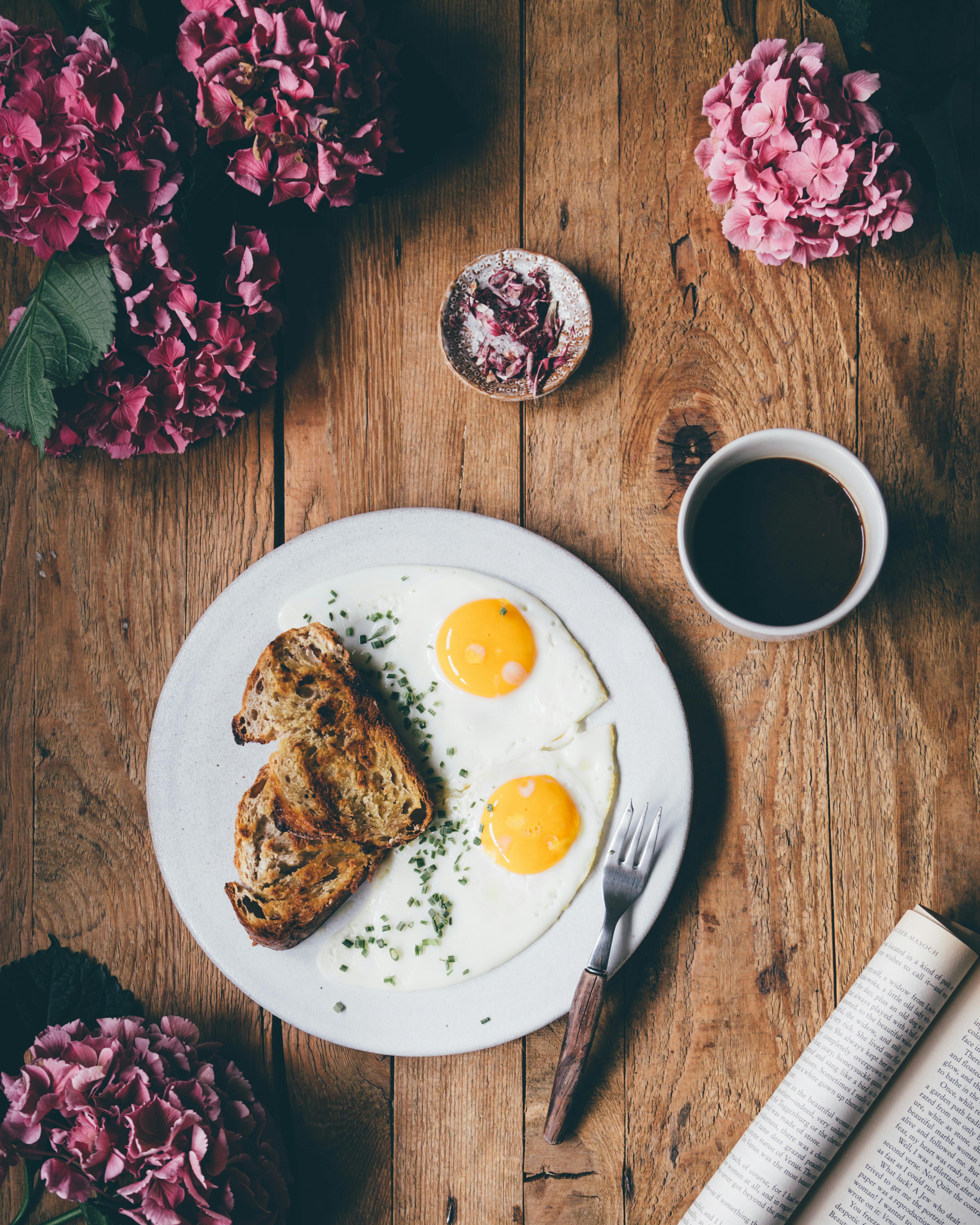 Top view flat lay of fried eggs with bread on white plate served on wooden table near cup of coffee and lush pink flowers