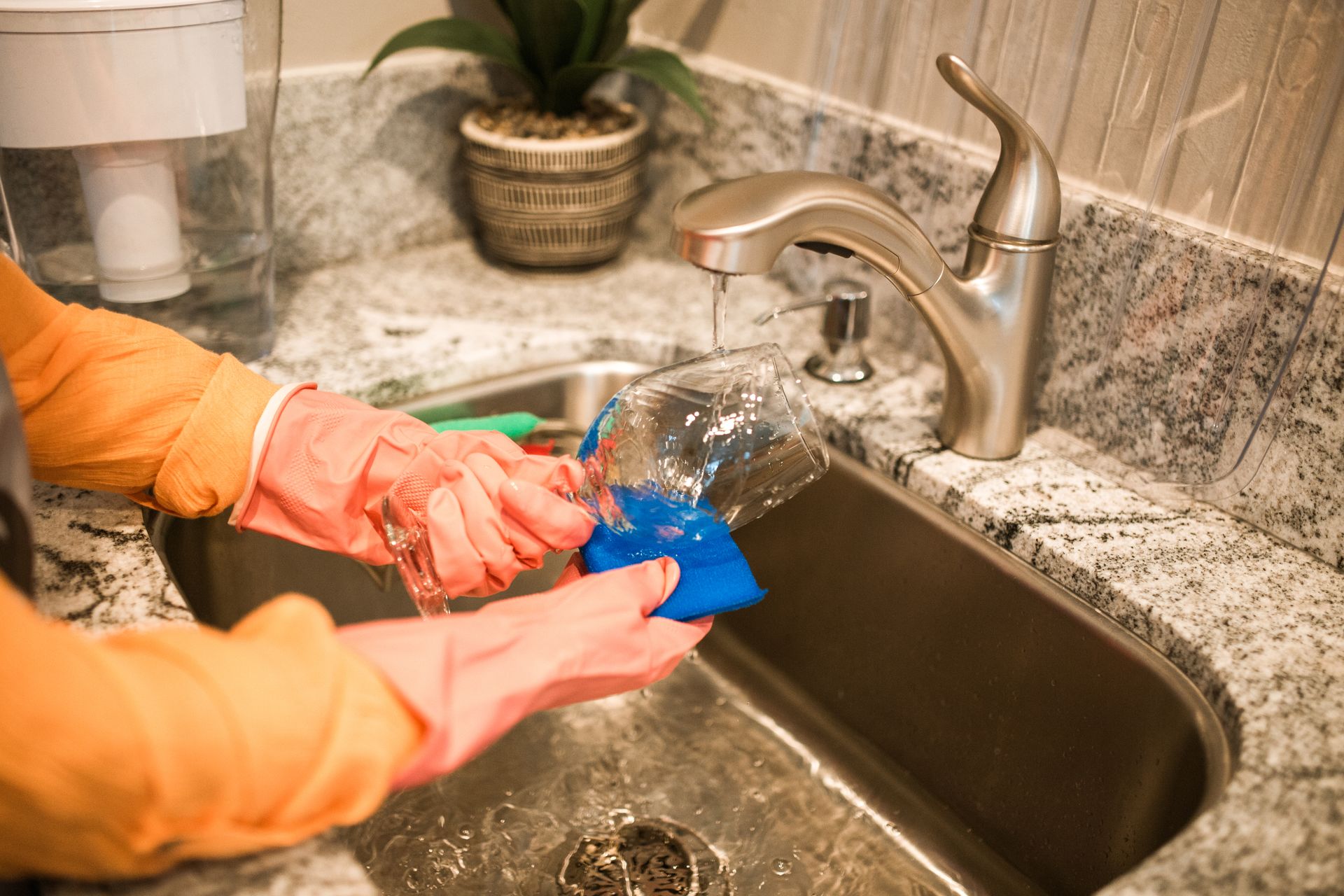 Close-up of hands wearing gloves washing a glass in a kitchen sink under a running faucet.