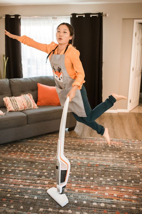 Free Asian woman joyfully vacuuming in mid-air, creating a playful home cleaning scene. Stock Photo