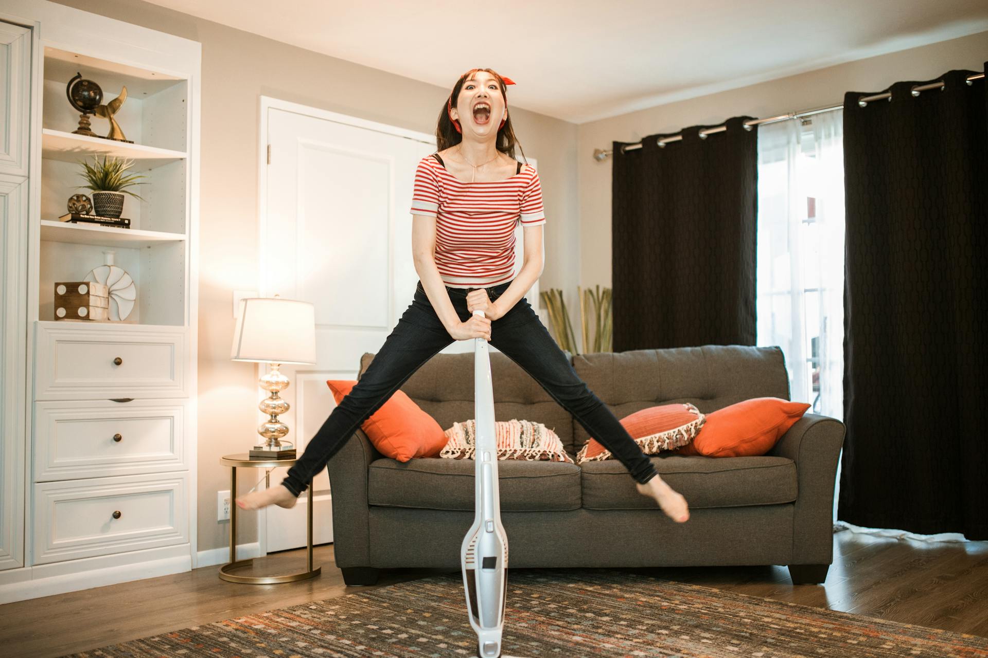 A joyful woman making cleaning fun by jumping with a vacuum in her living room.