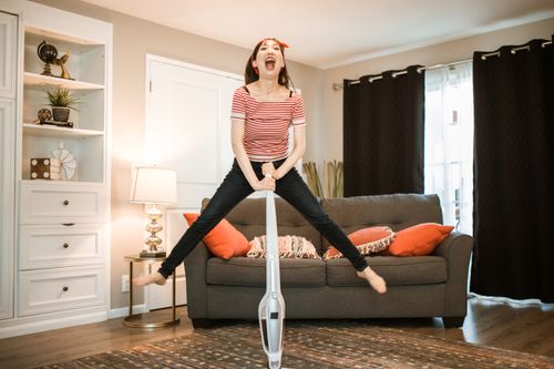Free A joyful woman making cleaning fun by jumping with a vacuum in her living room. Stock Photo