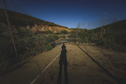 在山区，蓝天万里下，空荡荡的废弃运动场上，匿名者的影子映在受损的网状栅栏上