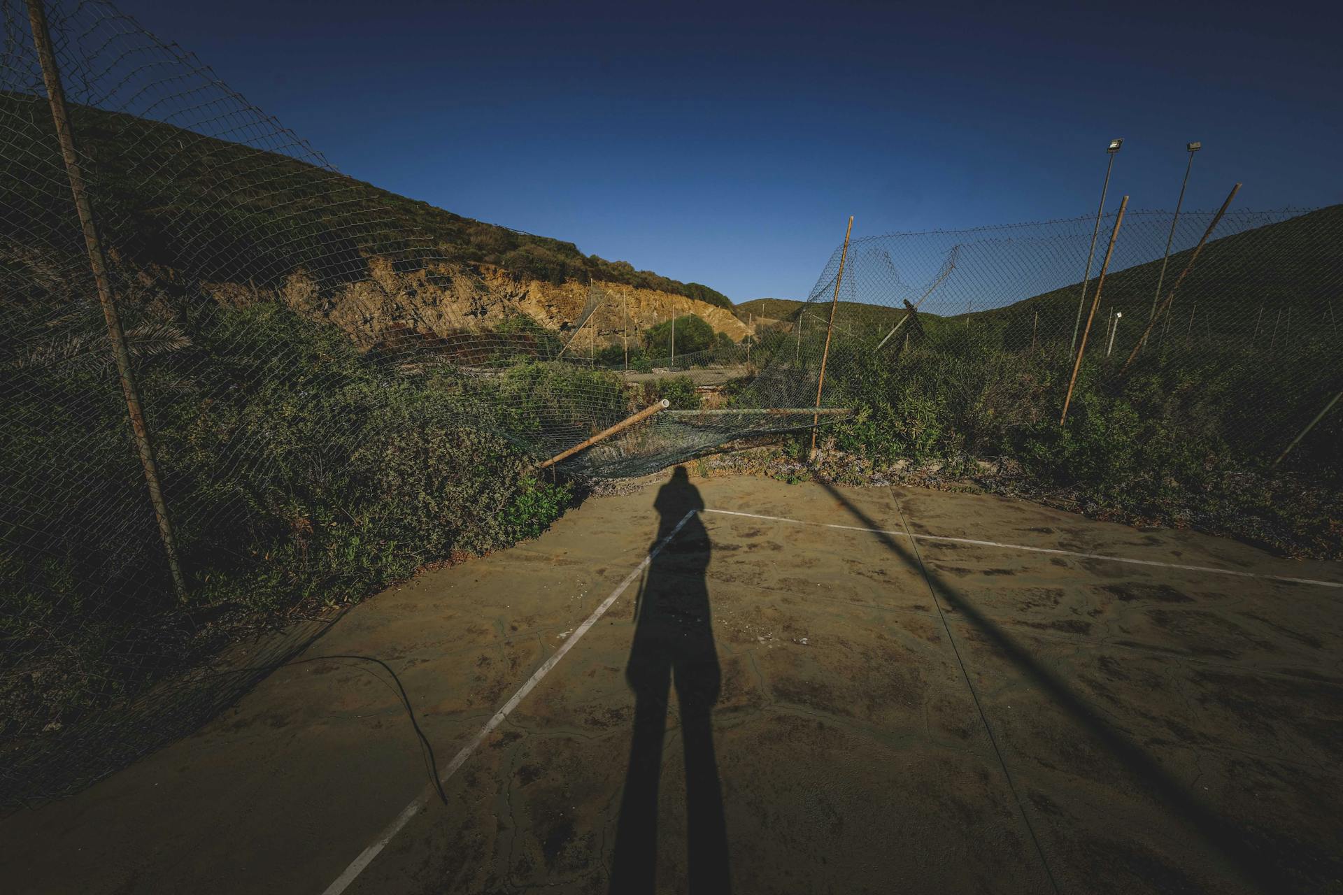 Shadow of anonymous person on empty abandoned sports ground with damaged mesh net fences located in mountainous terrain against cloudless blue sky