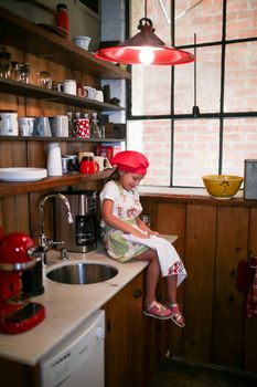 Cute young girl wearing a red chef hat, sitting by a kitchen sink playing pretend chef.