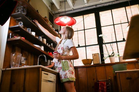 A woman in a floral apron arranging cups on a kitchen shelf, captured in warm indoor lighting.