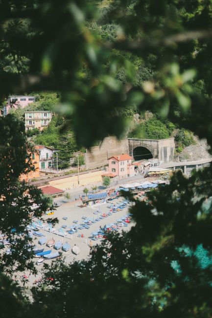 View through blurred greenery of old city beach filled with colorful boats and chaise lounges surrounded with houses and railway