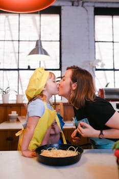A mother and daughter share a kiss while cooking pasta in a cozy kitchen.