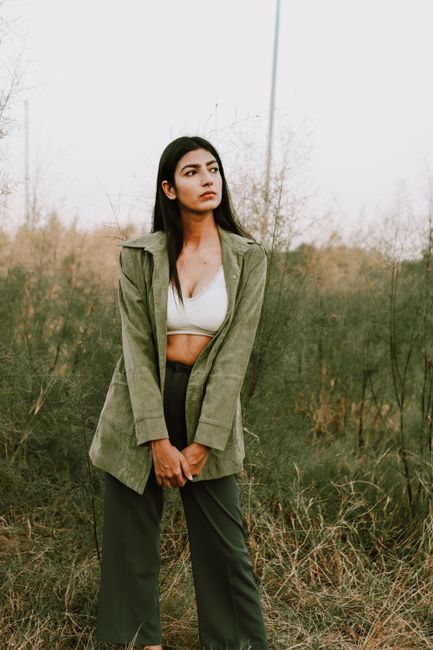 Pensive young ethnic lady with long straight hair in trendy crop top and blazer relaxing on grassy meadow in countryside against cloudy sky