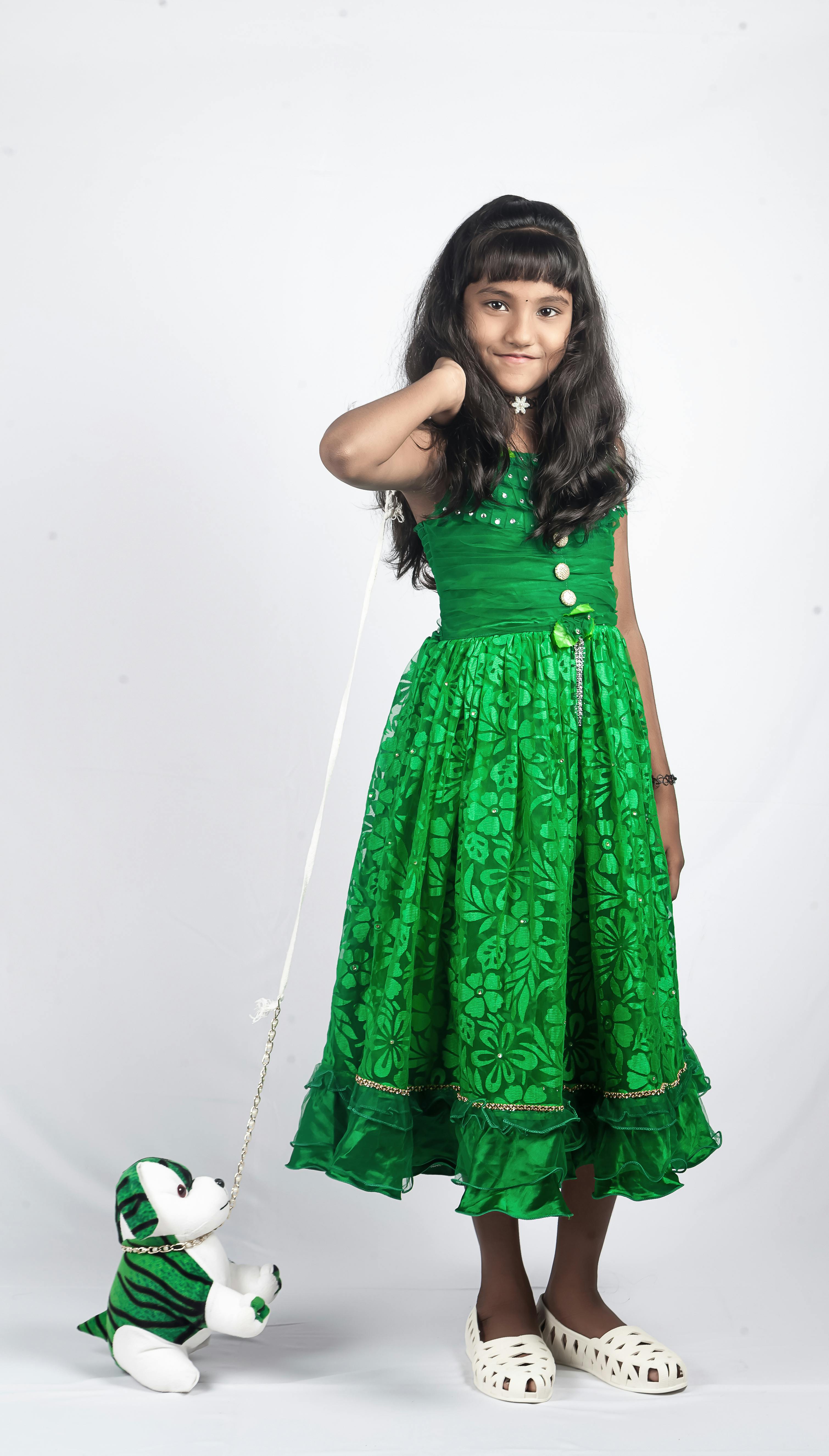 A cheerful young girl in a green dress poses with a stuffed toy dog on a white background.