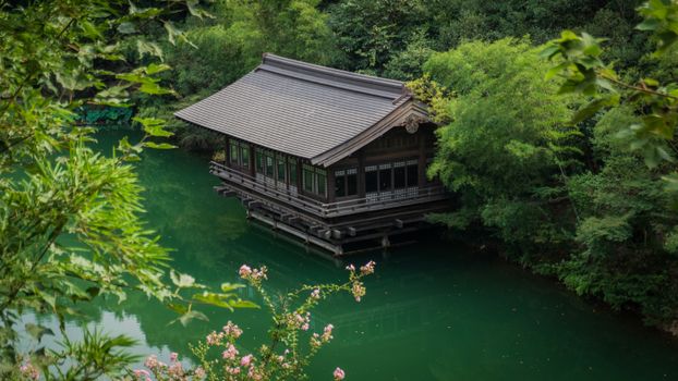 A tranquil view of a traditional Japanese pavilion over a serene green river surrounded by lush trees.