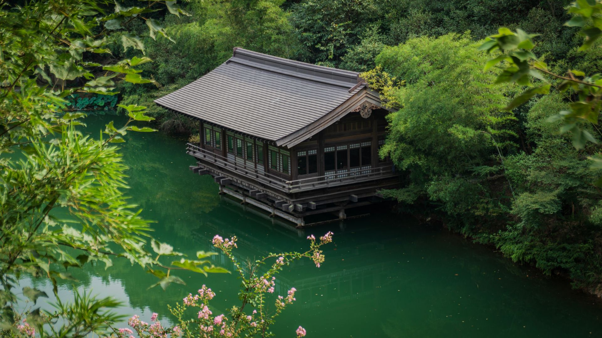 A tranquil view of a traditional Japanese pavilion over a serene green river surrounded by lush trees.
