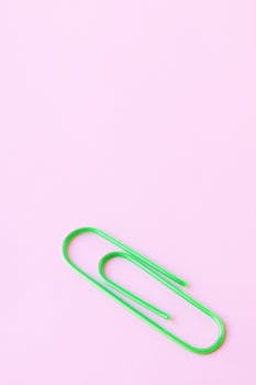 Close-up of a green paperclip on a pastel pink surface, perfect for stationary themes.