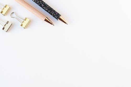 Top view of various brilliant pens and golden binder clips on straight white surface