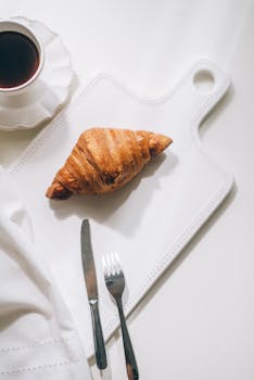 Stylish breakfast arrangement with a croissant on a cutting board and a cup of black coffee on a white background.