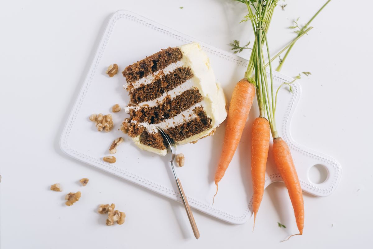 Slice of four-layer carrot cake with cream cheese frosting, walnuts and fresh carrots